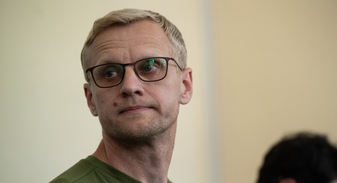 A man with short blondish hair and black-framed glasses looks directly at the camera with a neutral expression, wearing an olive green shirt against a beige wall background.