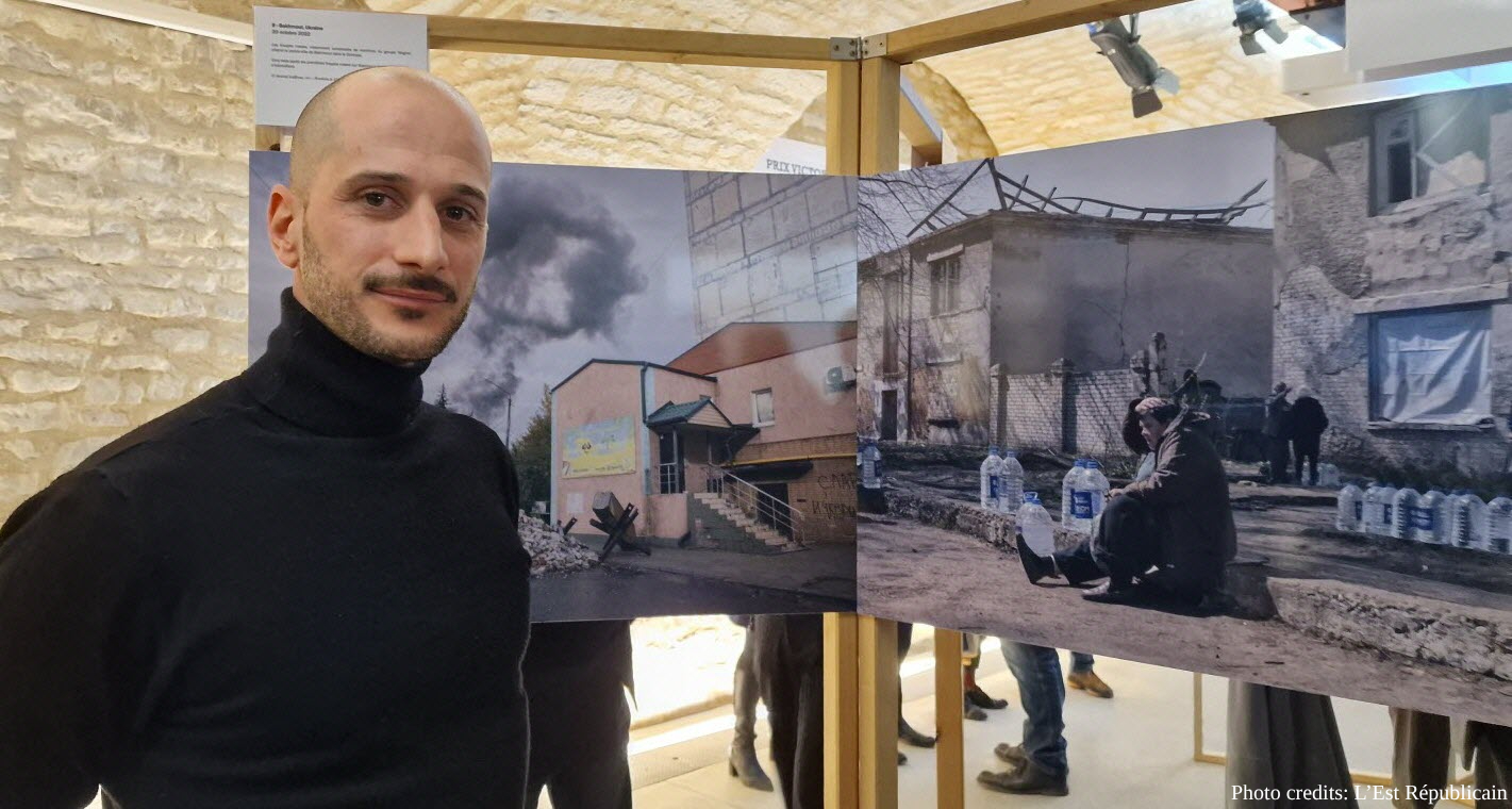 A man in a black turtleneck, Anthoni Lallican, stands beside large-format photographs displayed at an exhibition, showing scenes of war damage including a bombed building with rising smoke and people gathering near destroyed structures. The exhibition appears to document conflict aftermath, with the photographs mounted on wooden frames against a textured stone wall.