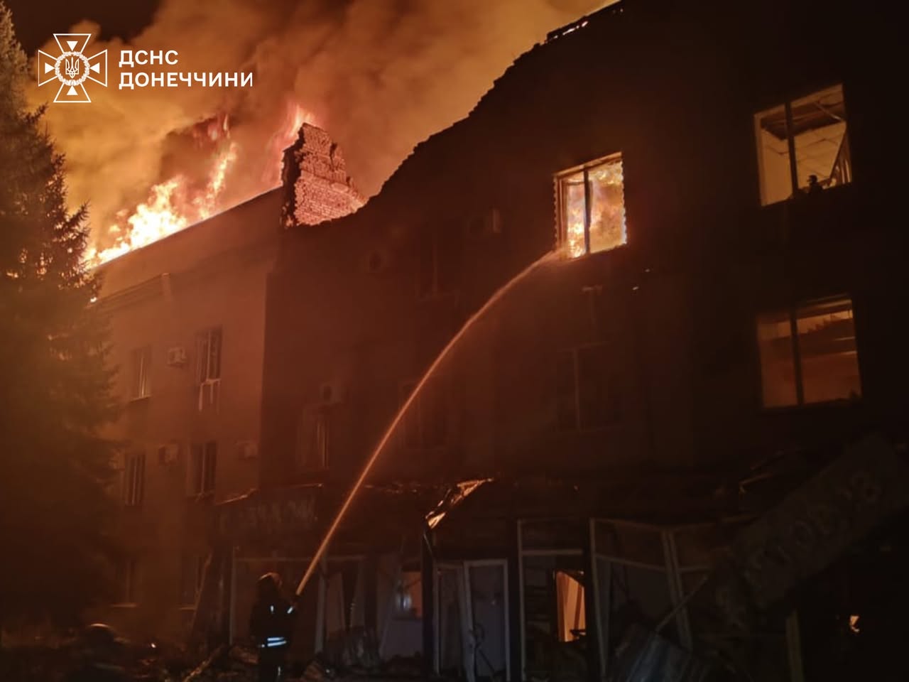 A multi-story residential building engulfed in intense flames at night, with fire consuming the upper floors and roof while firefighters direct water streams at the blaze from ground level. The structure shows significant damage with portions of the roof collapsed and windows illuminated by the fire's glow, captured in footage from the State Emergency Service of the Donetsk region.