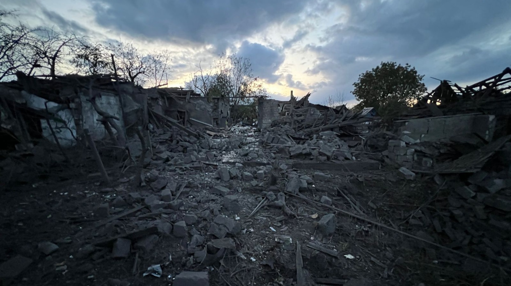 Destroyed residential buildings reduced to rubble and twisted metal beams under a dramatic cloudy sky at dusk, with bare and leafless trees visible in the background. The scene shows extensive structural damage with debris scattered across the ground and partially standing walls amid the devastation.