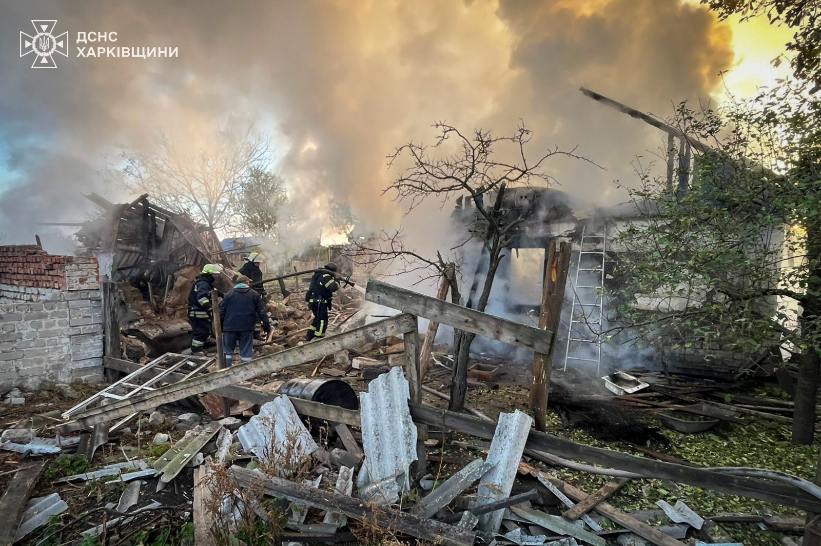Firefighters battle a blaze amid the destroyed remains of residential buildings, with thick smoke billowing from burning structures surrounded by rubble, bare trees and debris. The State Emergency Service of Kharkiv Region logo appears in the top left corner of this scene of destruction.