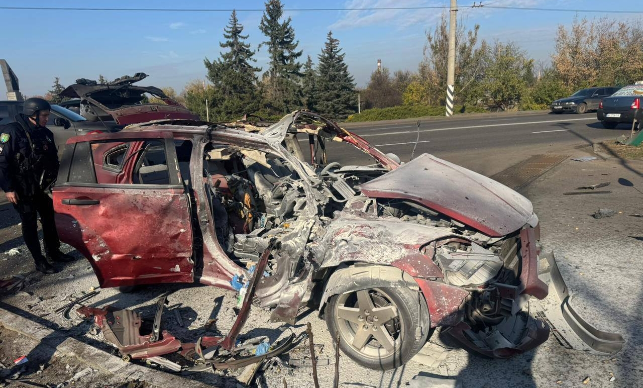 A severely destroyed red vehicle sits on a roadside with its roof completely torn off, front end crushed, and interior gutted, while police officers investigate the scene near evergreen trees under a clear sky. The wreckage shows extensive structural damage with debris scattered around the collision site on what appears to be a rural highway.