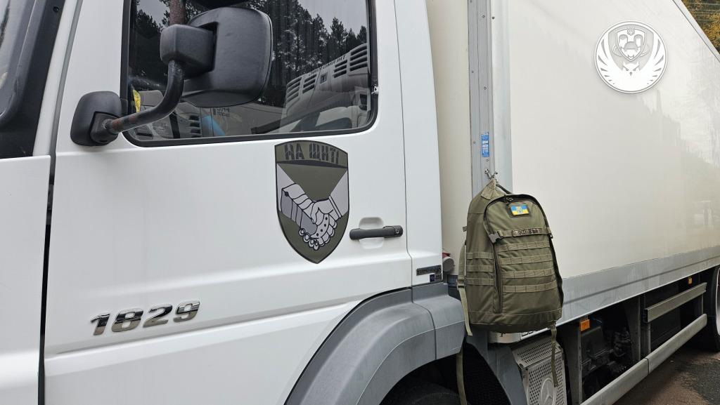 A white delivery truck with a shield emblem featuring clasped hands and Ukrainian text on the cab door, with an olive-green military backpack bearing a Ukrainian flag patch hanging from the door handle. The truck appears to be part of a humanitarian logistics operation.