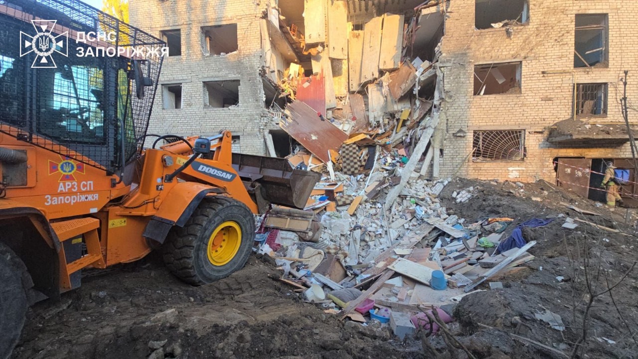 An orange Doosan front-end loader clears debris from a heavily damaged multi-story residential building in Zaporizhzhia, with collapsed walls and rubble piled in front of the structure. The image bears the State Emergency Service of Ukraine logo.