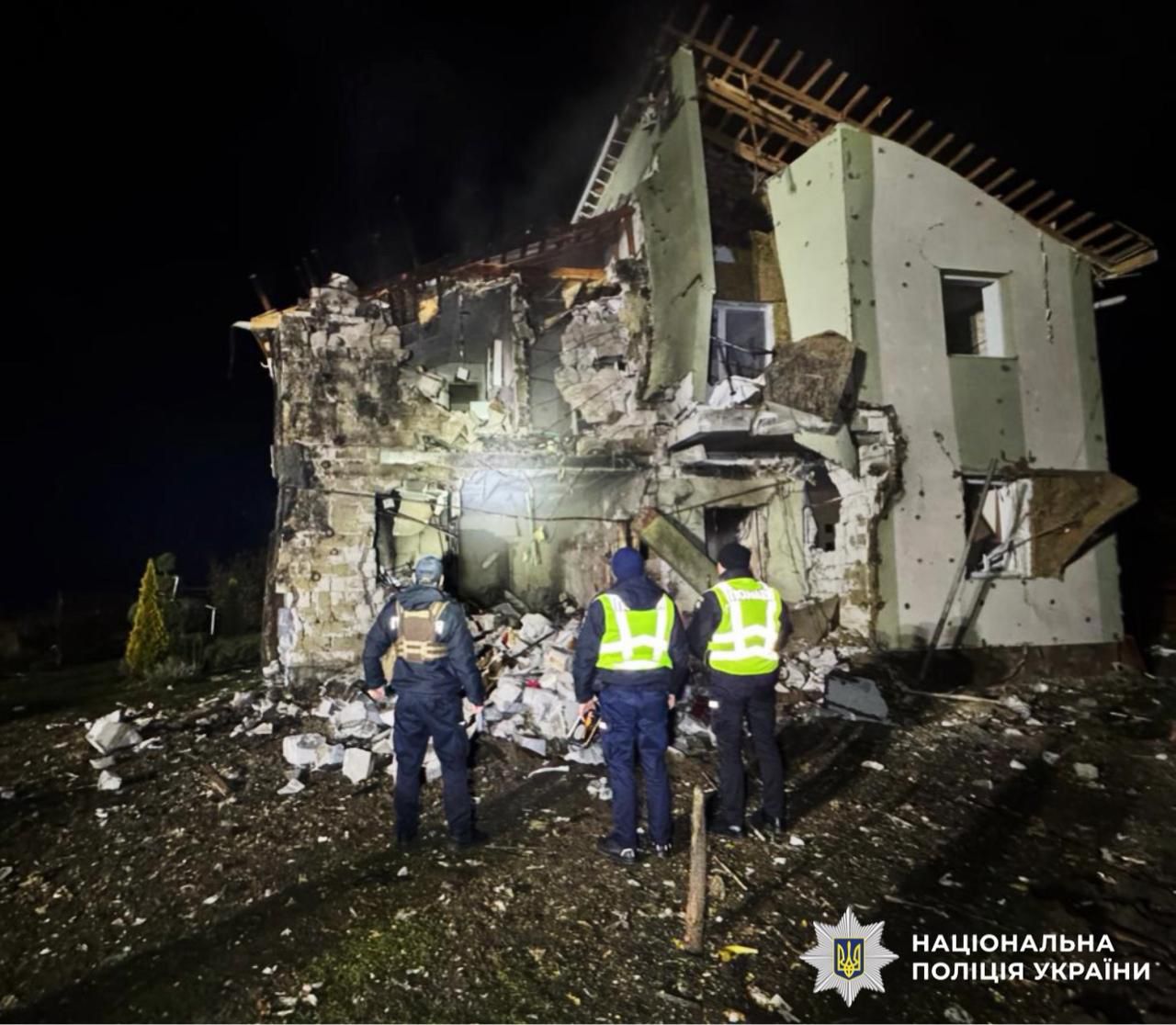 Three emergency responders in reflective vests examine a severely damaged two-story residential building at night, with debris scattered across the ground and walls partially collapsed. The image is marked with the National Police of Ukraine logo.