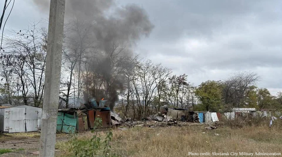 Dark smoke rises from burning structures and debris in an overgrown lot with bare trees, small sheds and scattered wreckage visible across the landscape under an overcast sky. A concrete utility pole stands in the foreground, with photo credit attributed to Sloviansk City Military Administration.