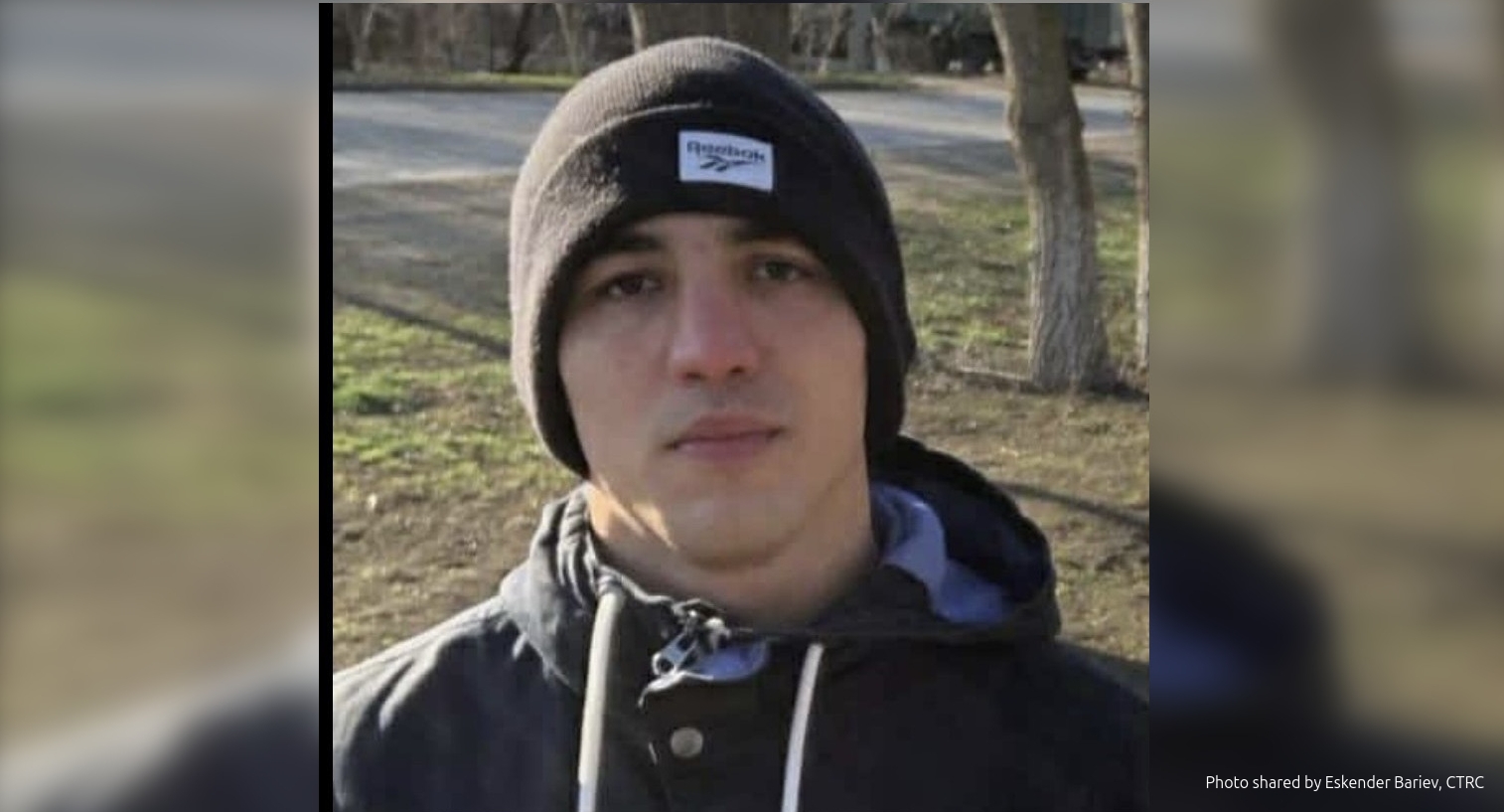 A young man, a Crimean Tatar Mamut Belyalov, wearing a knit beanie with a label and a dark jacket takes a selfie outdoors in a park setting with bare trees and a road visible in the background. The photo appears to be taken during winter or early spring with natural daylight.