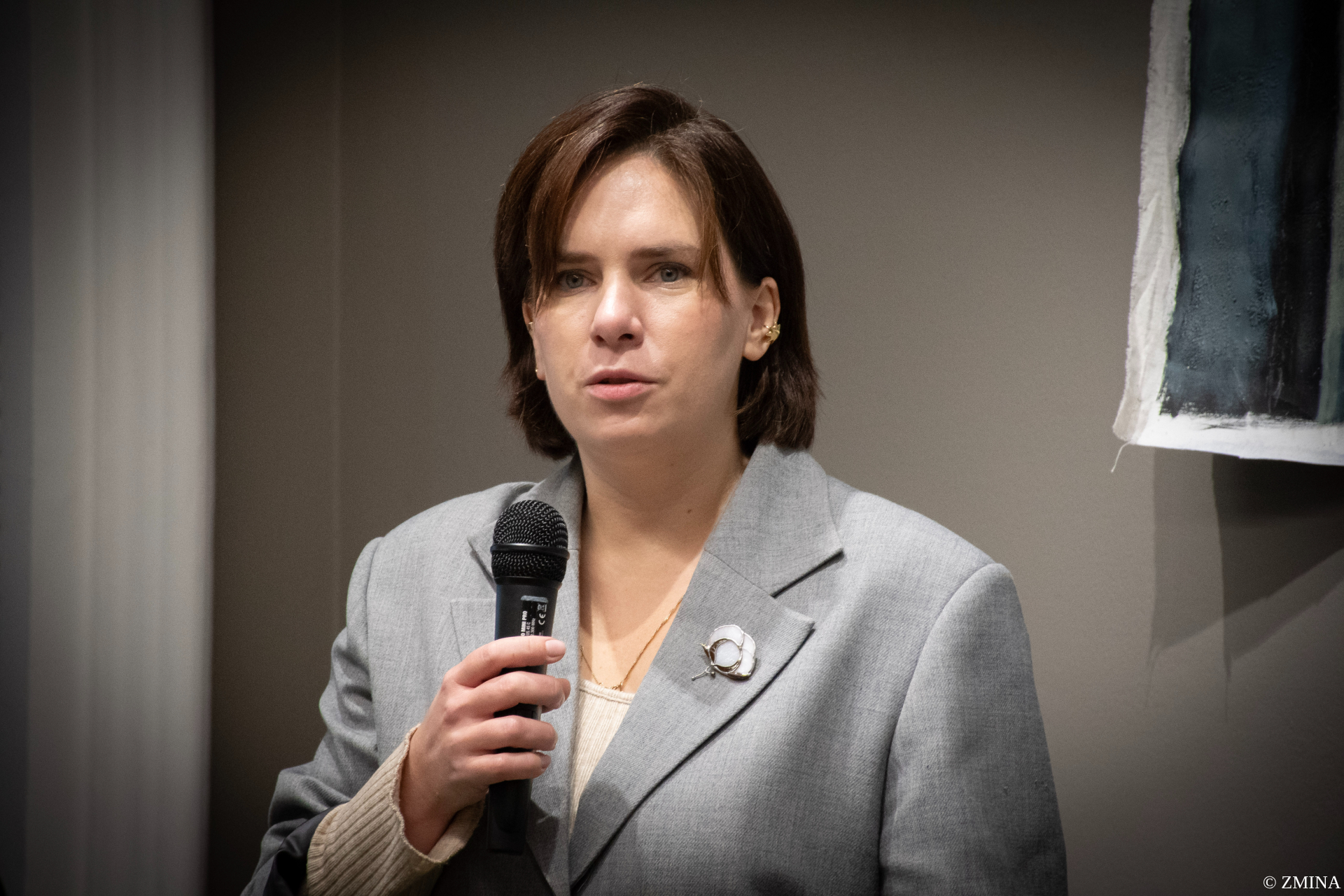 A woman with shoulder-length brown hair wearing a light gray blazer and beige top speaks into a microphone, with a circular pin attached to her lapel. She appears to be addressing an audience in a professional setting with neutral-toned walls visible in the background.Retry