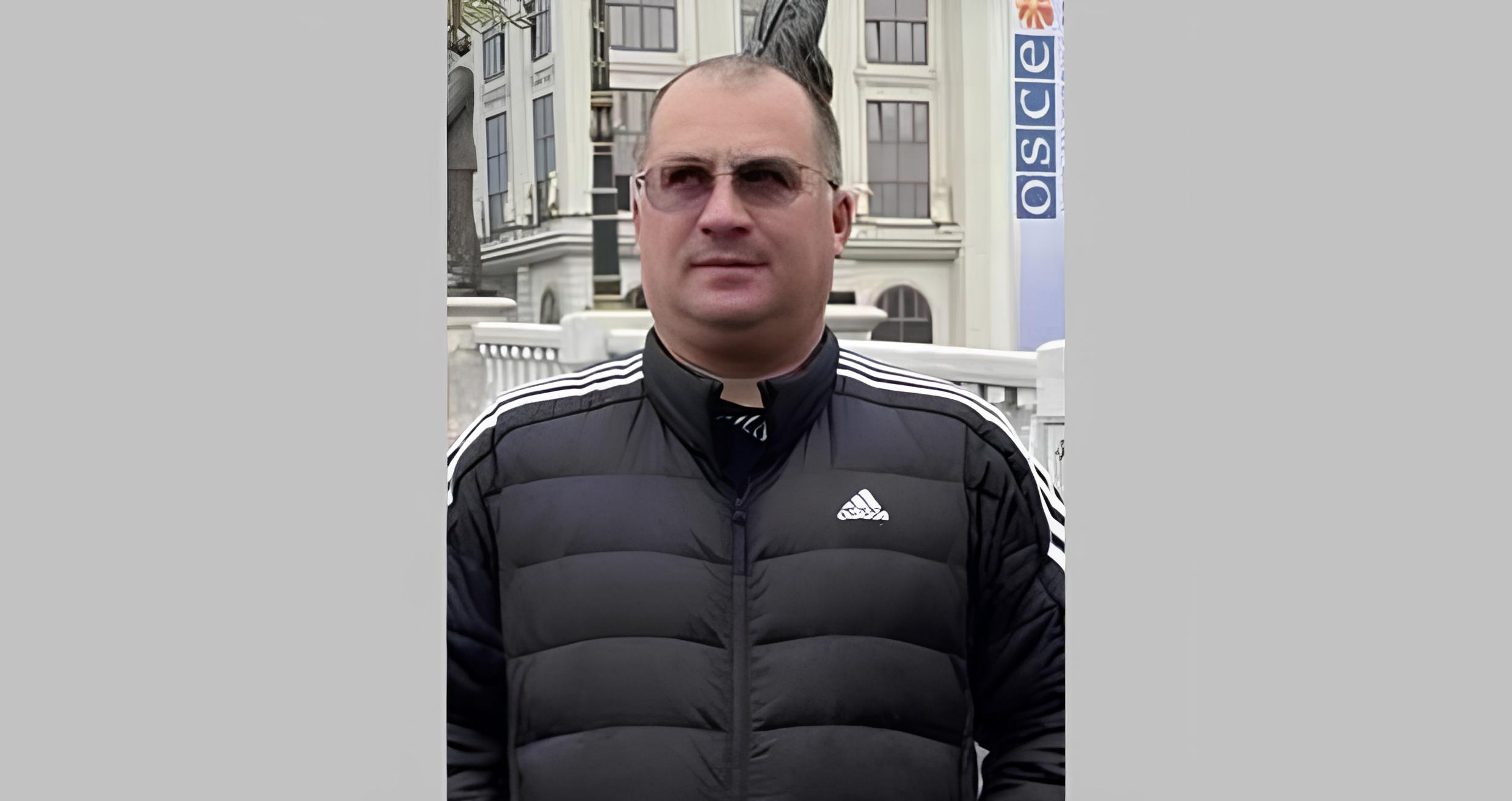 A man wearing sunglasses and a black Adidas puffer vest with white stripes stands in front of a building with white columns and an OSCE banner visible in the background. The setting appears to be at an official or institutional location with classical European architecture.