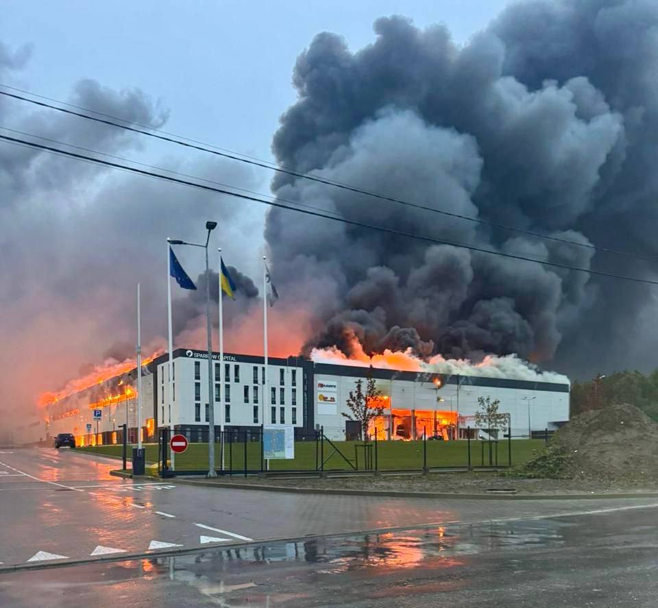 A modern commercial building engulfed in flames with massive plumes of black and gray smoke rising into the sky, as orange fire consumes the structure's roof and sides. The Ukrainian flag flies among flagpoles in front of the burning building on a wet street, with power lines visible overhead.