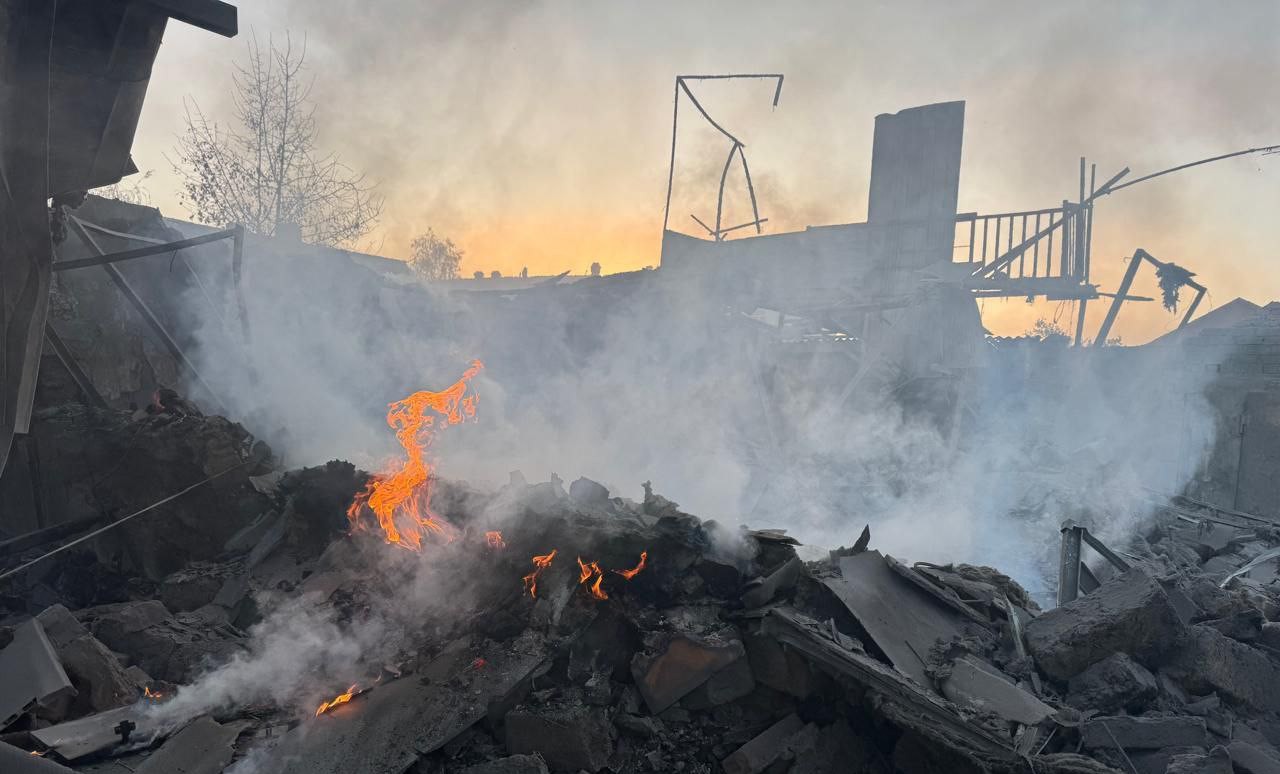 Flames and smoke rise from a pile of rubble and debris where a structure once stood, with skeletal remains of walls and framework visible against a smoke-filled sky. The destruction shows scattered concrete blocks, twisted metal, and charred materials still smoldering in the aftermath of what appears to be a building after Russian shelling.