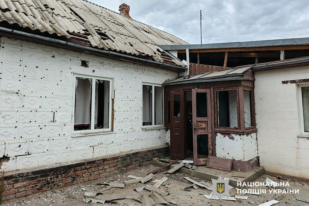 A heavily damaged residential house with white walls pockmarked by numerous impact holes, a partially collapsed corrugated roof, broken windows, and debris scattered around the entrance. The structure shows signs of conflict-related damage, with the National Police of Ukraine logo visible in the corner of the documentation photograph.