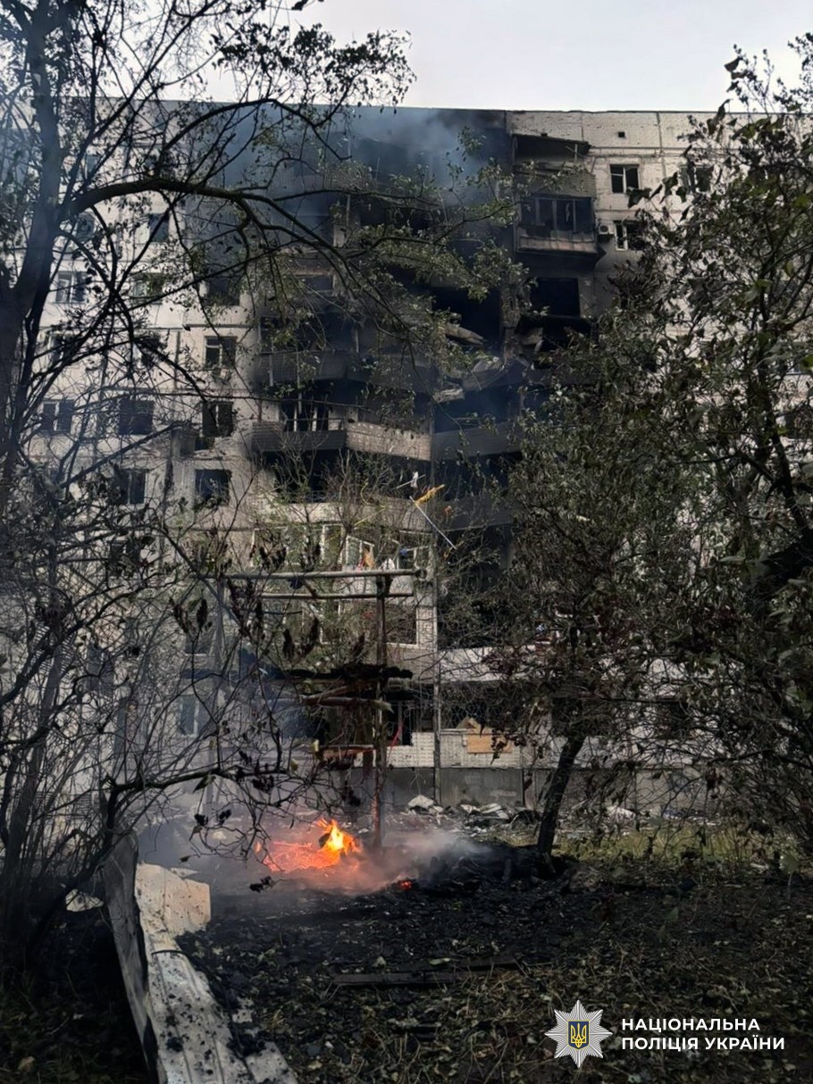 A severely damaged multi-story residential building with blown-out windows and structural damage stands behind bare trees, with debris scattered on the ground and small fires burning in the courtyard area. The scene shows the aftermath of destruction, documented by the National Police of Ukraine as indicated by their logo in the bottom corner.