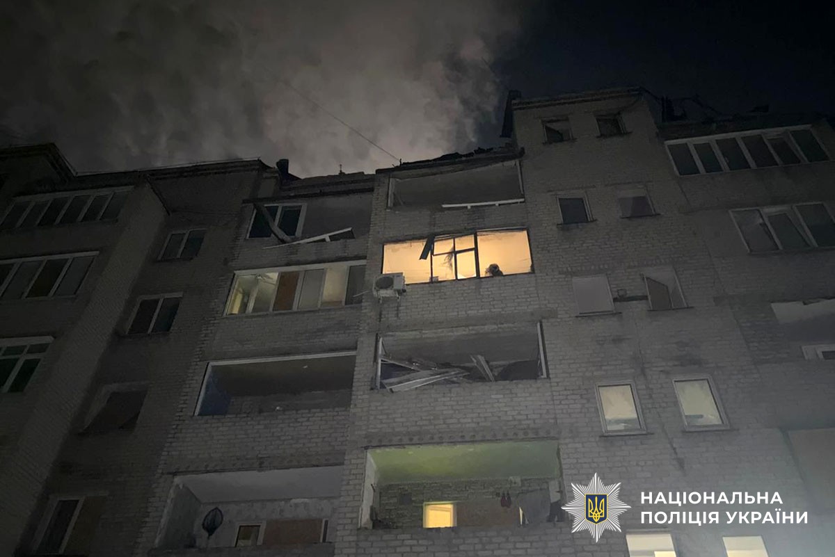 A damaged residential apartment building photographed at dusk or night, with smoke rising from the upper floors and visible structural damage including blown-out windows and broken balconies. A few illuminated windows glow yellow against the gray facade, with the National Police of Ukraine logo visible in the lower right corner.