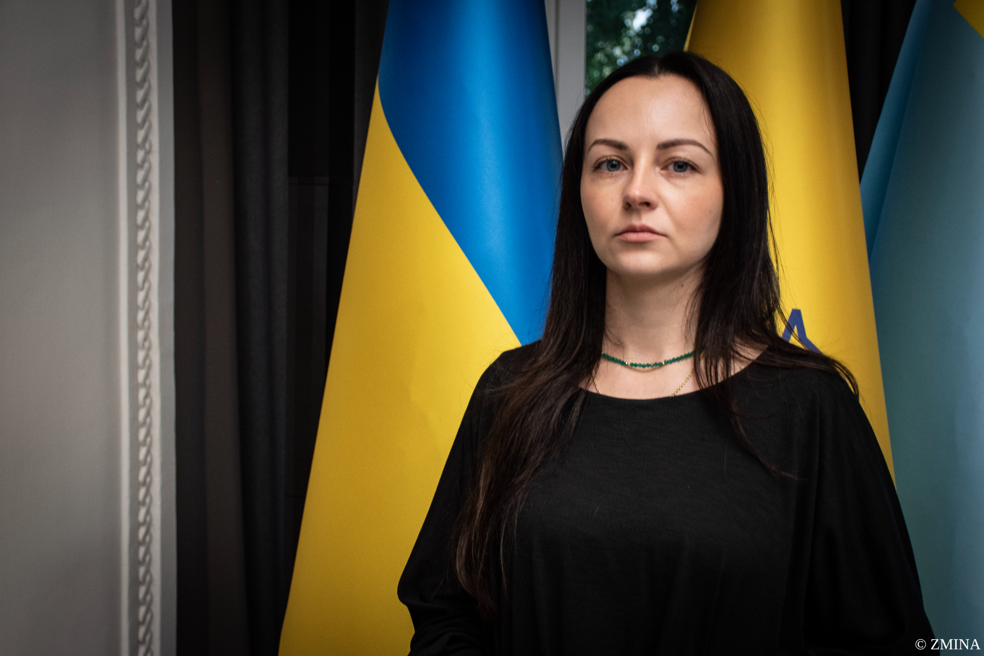 A woman with long dark hair wearing a black top and green beaded necklace stands in front of Ukrainian flags featuring blue and yellow stripes. She gazes directly at the camera with a serious expression in what appears to be an official or formal setting.