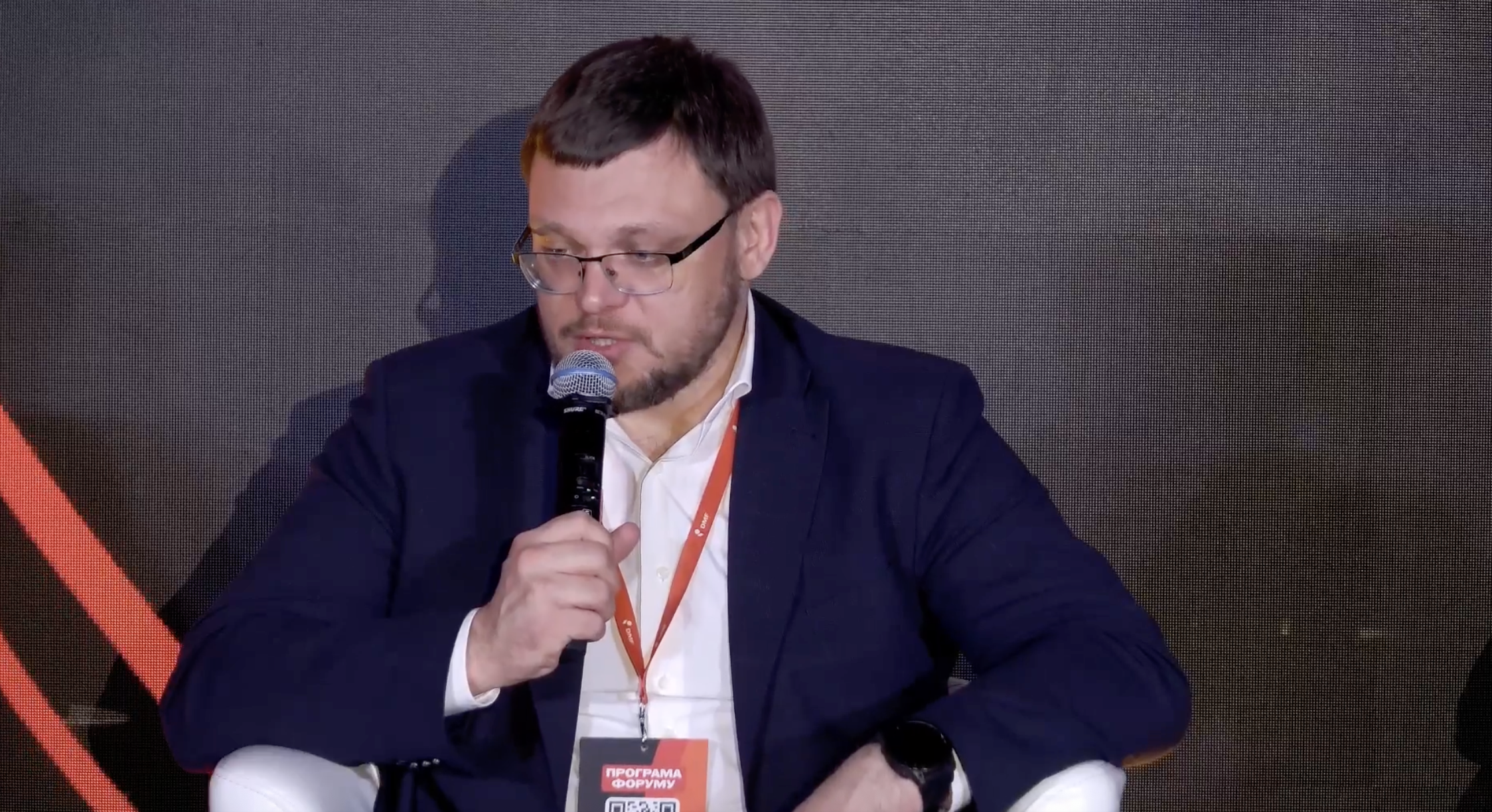 A man wearing glasses and a dark blue suit jacket over a white shirt speaks into a microphone while wearing a red lanyard with conference credentials. He is participating in a panel discussion at the Donbas Media Forum.