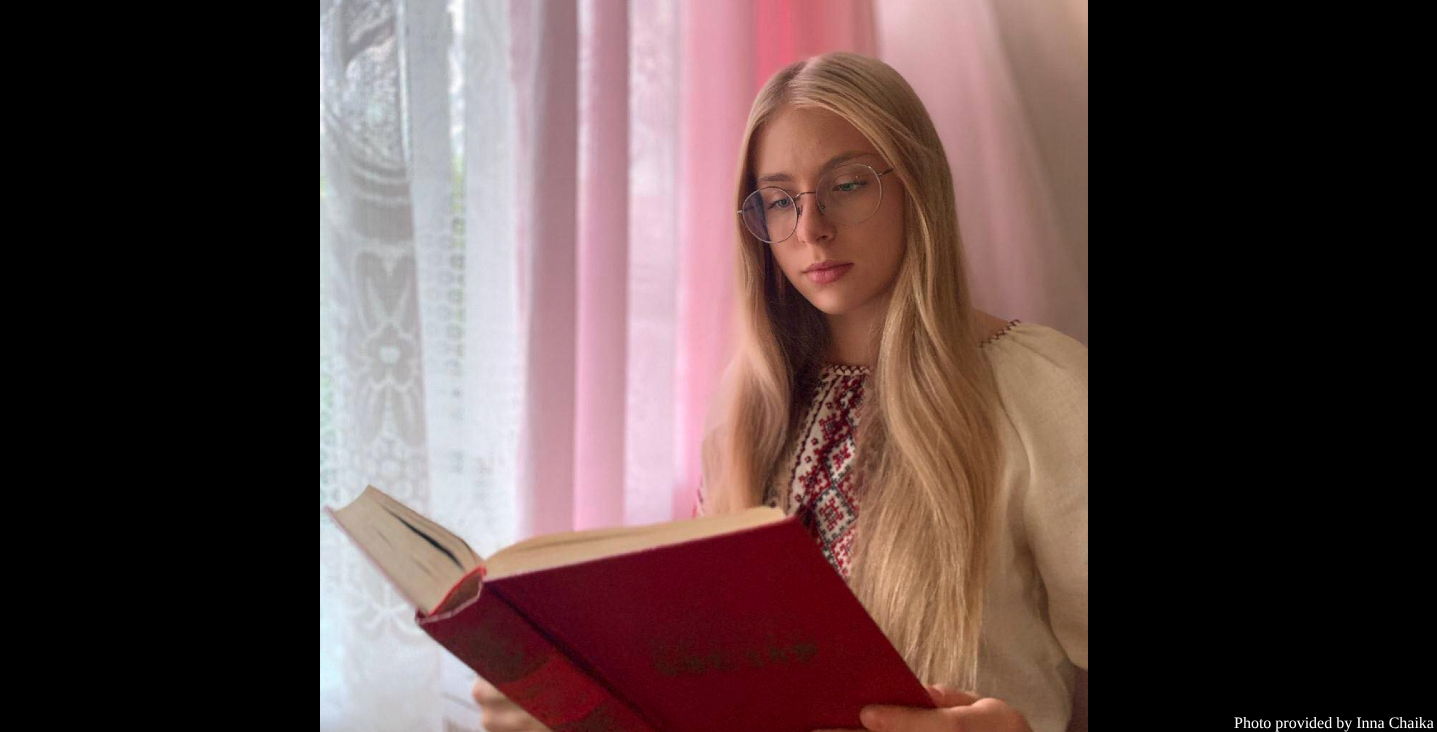 A young woman with long blonde hair and round glasses sits reading a red book while wearing a cream-colored embroidered blouse with traditional Ukrainian folk detailing. Soft natural light filters through sheer pink curtains behind her, creating a serene, intimate reading atmosphere.