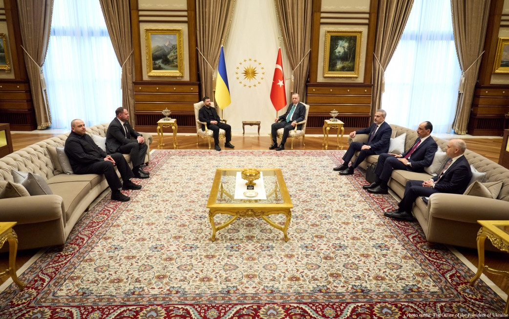 Government officials meet in an ornate reception room with beige tufted sofas arranged around a gold-framed coffee table on an elaborate Persian rug, with Ukrainian, Turkish, and presidential flags displayed between tall curtained windows and framed paintings on wood-paneled walls.