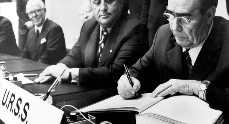 A diplomat in glasses and a dark suit signs an open document at a conference table, with a "U.R.S.S." nameplate visible and other formally dressed officials seated in the background. The black-and-white photograph captures what appears to be an official treaty or agreement signing ceremony.
