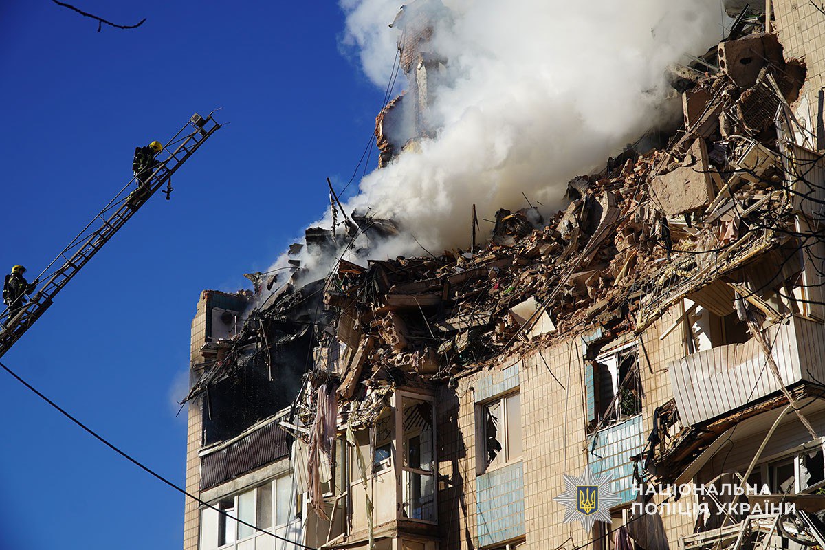 A firefighter on an extended ladder battles flames and thick smoke billowing from the severely damaged upper floors of a multi-story residential building in the city of Ternopil, Ukraine, with exposed interiors and collapsed sections. The catastrophic structural damage shows blown-out walls, dangling debris and twisted metal against a clear blue sky as emergency crews work to contain the fire.