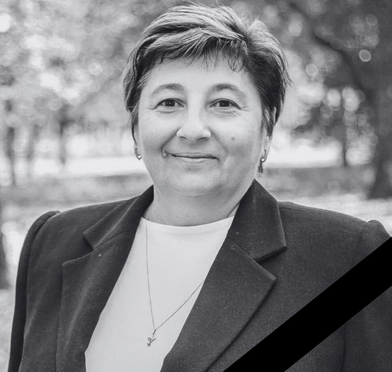 A black and white memorial portrait of Kherson resident shows a middle-aged woman with short hair wearing a dark blazer over a white top with a pendant necklace, photographed outdoors with a blurred background. A black ribbon crosses the lower right corner, indicating this is a commemorative image.