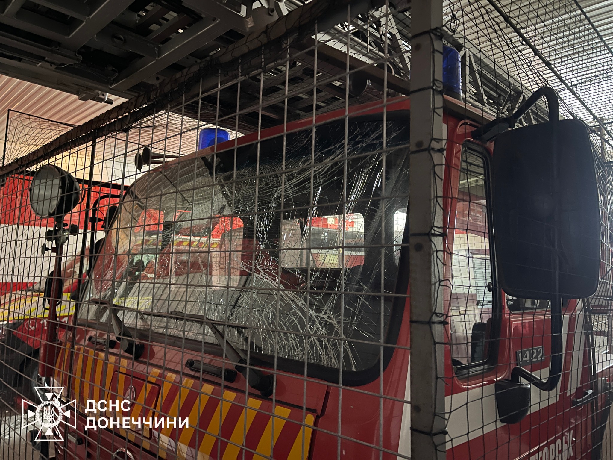 A fire truck with shattered windshield and damaged front section sits inside a protective metal cage structure within what appears to be a fire station garage. The State Emergency Service of Donetsk region logo appears in the bottom left corner of the image documenting the damaged emergency vehicle.