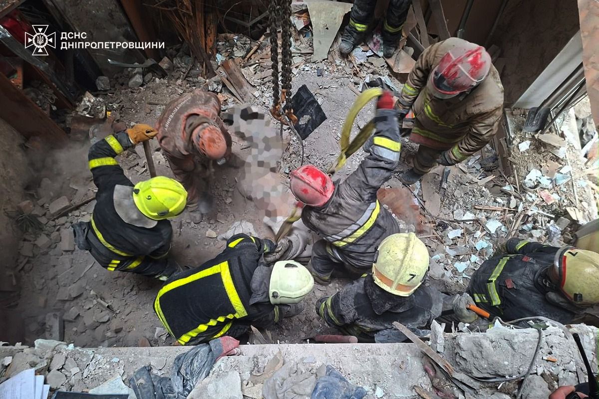 Emergency responders in protective gear and helmets work to extract a victim from rubble and debris in a destroyed building, with the State Emergency Service of Dnipropetrovsk region logo visible in the upper left corner. The rescue team, wearing yellow and red helmets, carefully maneuvers through concrete fragments and twisted metal to reach the person beneath the collapsed structure.
