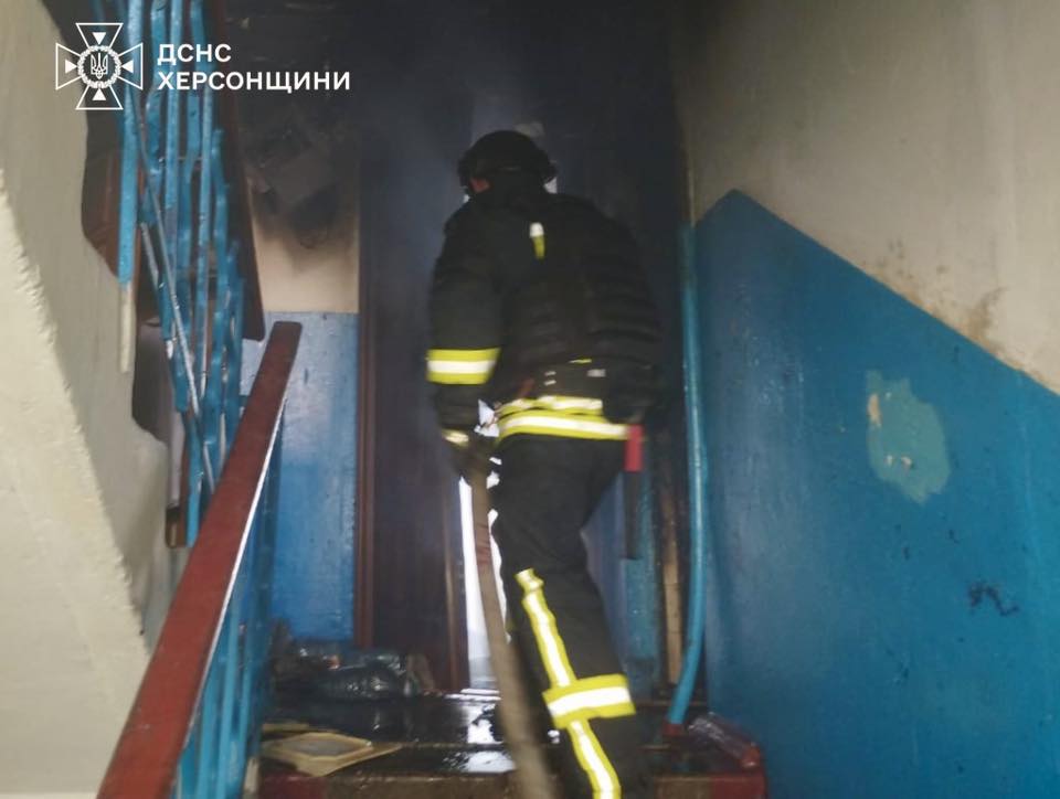 A firefighter in protective gear with reflective yellow stripes navigate through a smoke-filled apartment building stairwell with blue railings and worn walls. The State Emergency Service of Kherson region logo appears in the top left corner of the image documenting the emergency response.