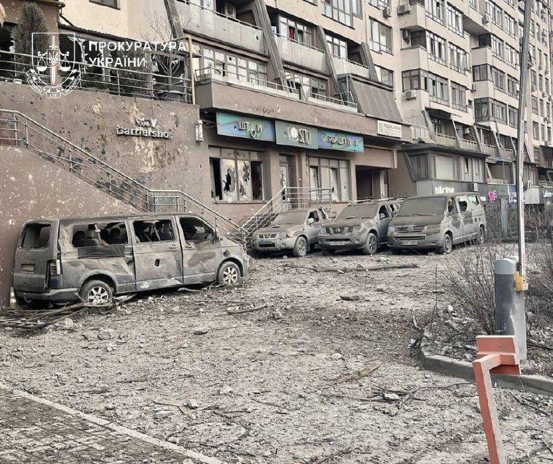 Multiple damaged vehicles sit in front of a shrapnel-scarred residential building with broken windows and damaged storefronts, debris covering the ground in this daytime scene marked with the Office of the Prosecutor General of Ukraine logo.