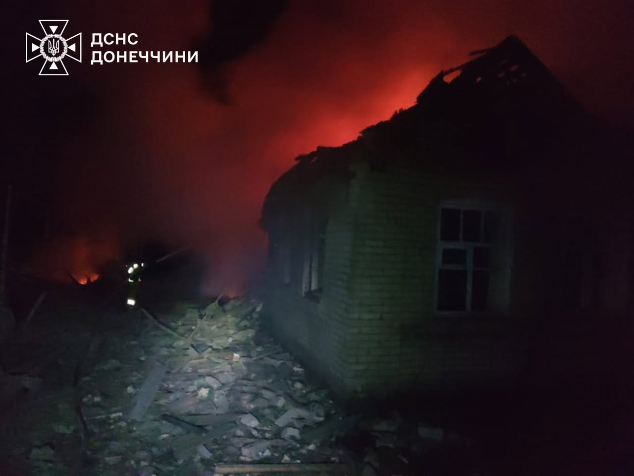Emergency responders work near a burning residential building at night, with intense orange flames and smoke visible against the dark sky. The damaged structure shows a collapsed roof and scattered debris, while firefighters with reflective gear are silhouetted in the foreground.