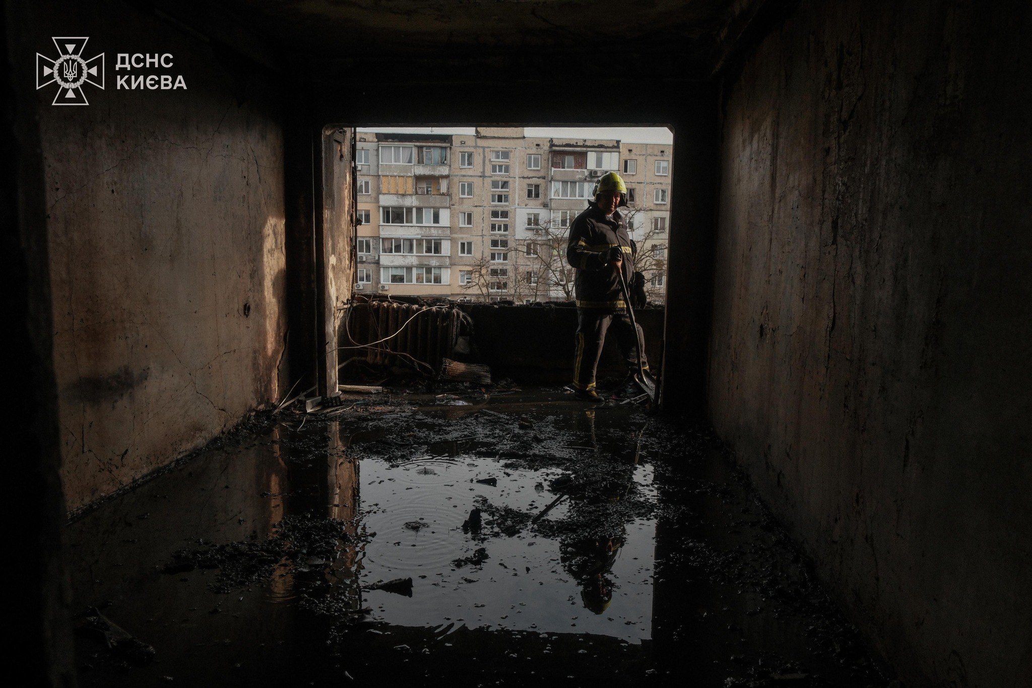 A firefighter in protective gear stands in a severely fire-damaged room with charred floors and walls, silhouetted against a window opening that reveals a multi-story residential building. The Kyiv State Emergency Service logo appears in the upper left corner.