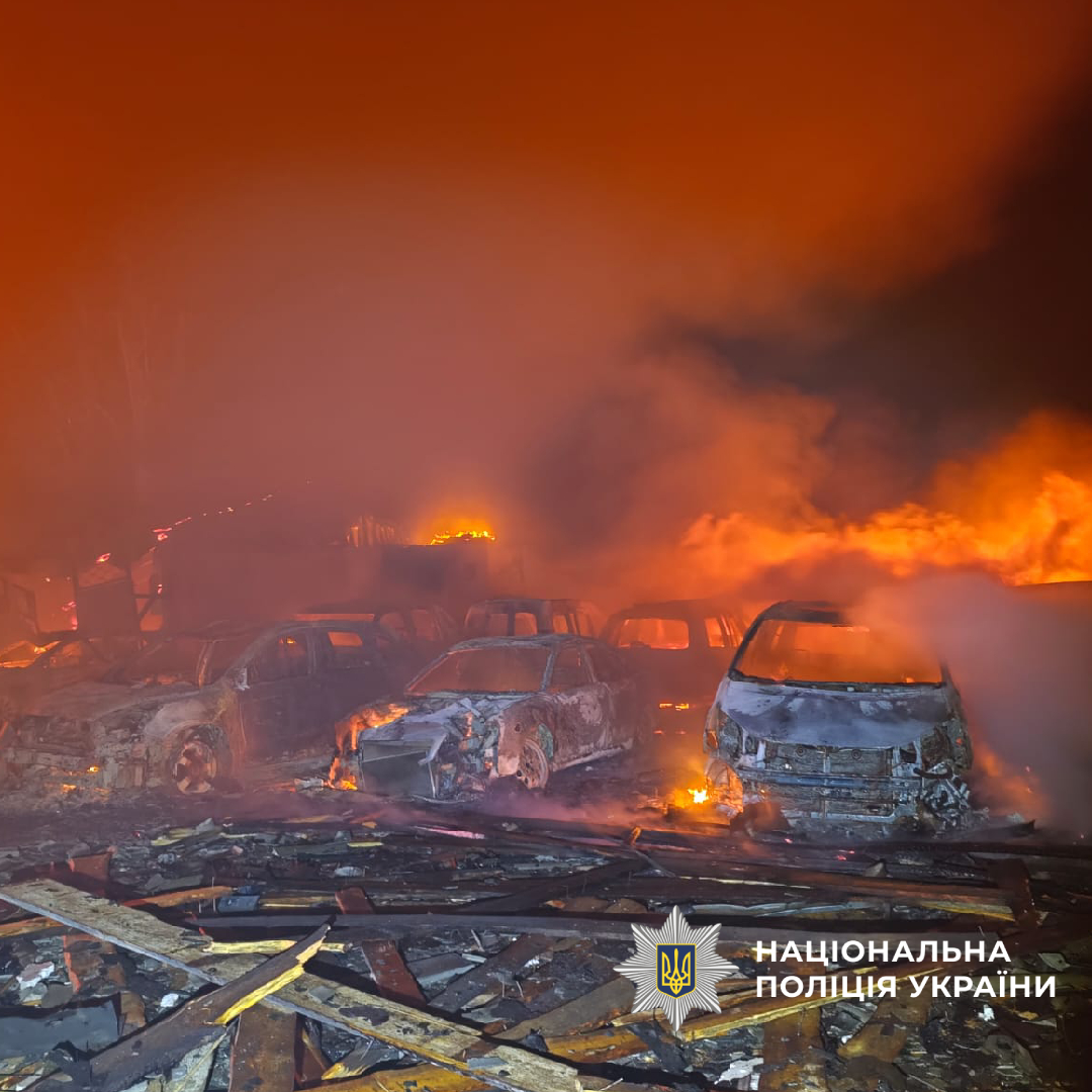 Multiple burned-out vehicles engulfed in flames and thick smoke with emergency responders visible in the background amid scattered debris and destruction. The National Police of Ukraine logo appears in the bottom right corner of this documentation of the catastrophic scene.
