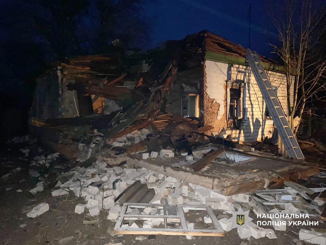 A severely damaged single-story house at dusk shows a collapsed roof and walls with extensive debris scattered across the ground, including white insulation pieces and wooden fragments. A ladder leans against the partially standing yellow wall, illuminated by interior lighting visible through damaged windows.