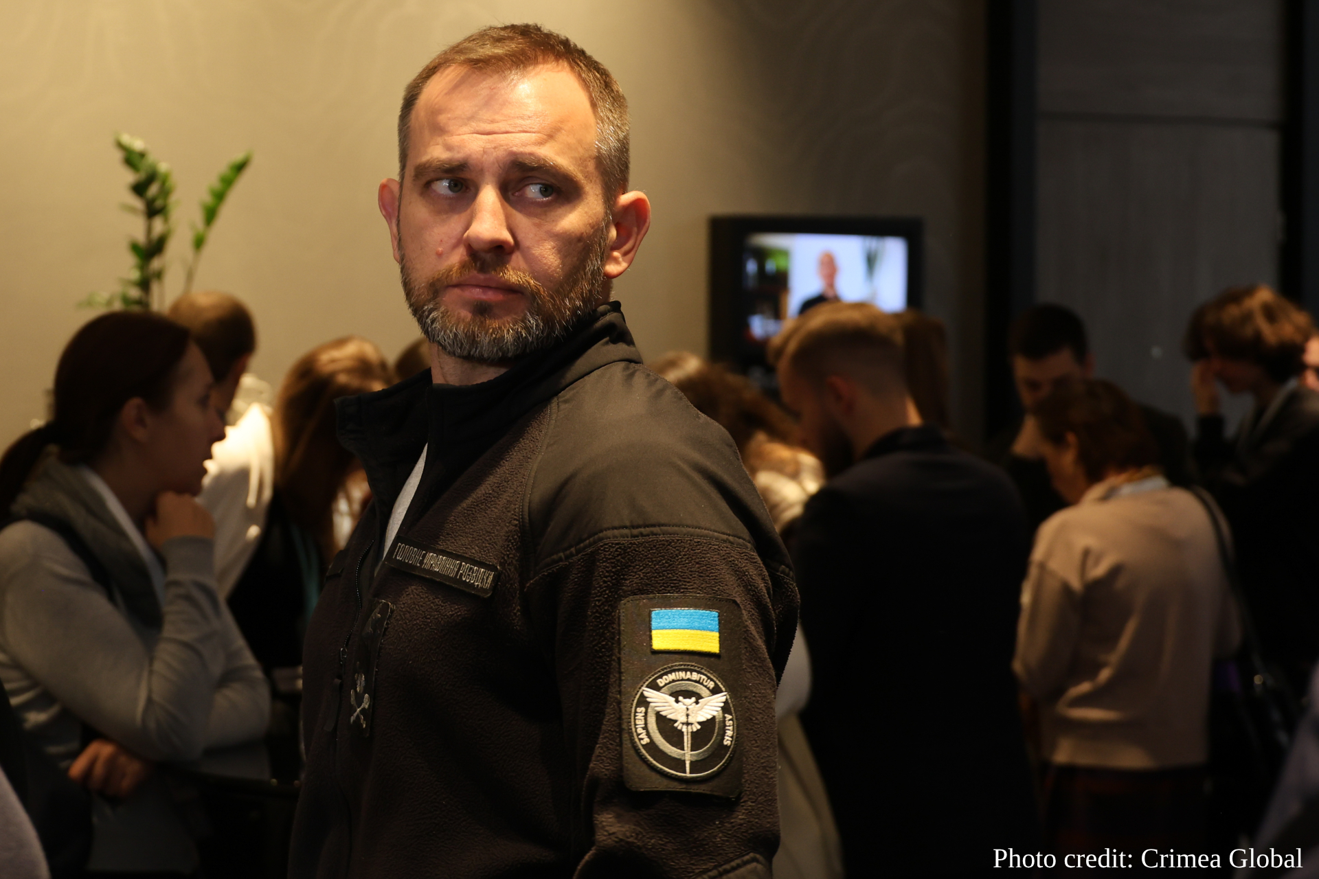 A handsome man, Dmytro Usov, in a dark tactical vest bearing a Ukrainian flag patch and military insignia looks directly at the camera while standing in a crowded indoor space with blurred attendees and a plant visible in the background.