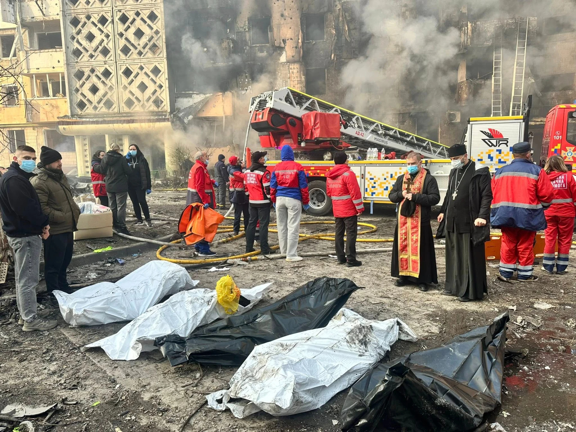 Bodies in black and white packing lie on the ground in the foreground as emergency responders in red uniforms, civilians, and an Ukrainian Orthodox priest gather near fire trucks and ladder equipment at a severely damaged, smoking apartment building in Ternopil after Russian missile. The scene shows active firefighting operations with visible fire hoses on the ground and smoke billowing from the blackened, damaged facade with blown-out windows.
