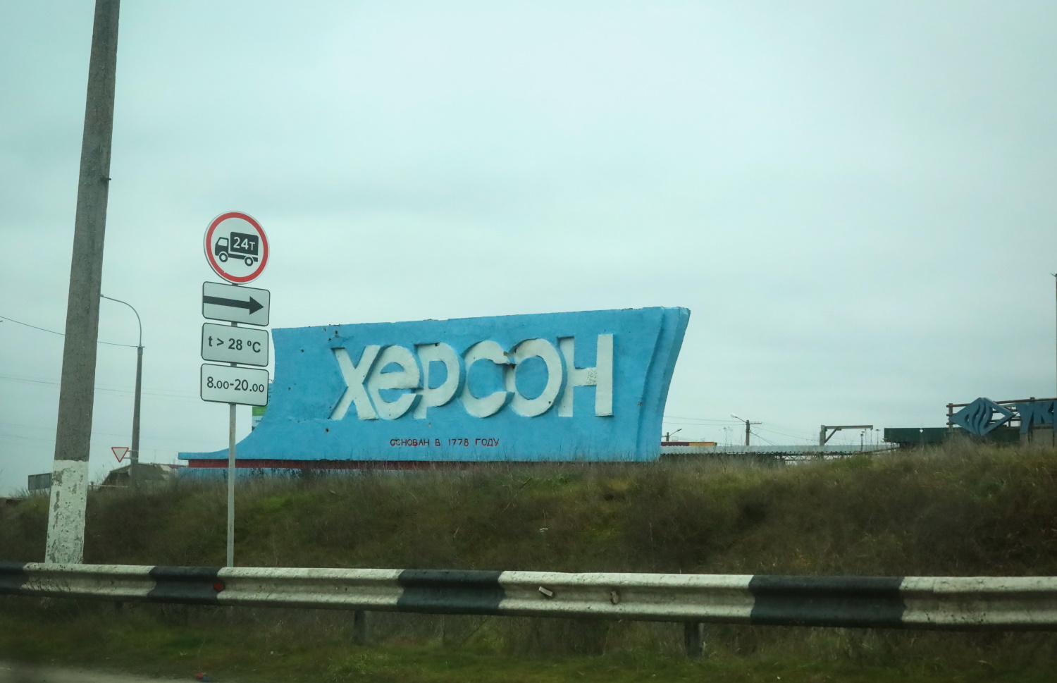 A weathered blue city entrance sign reading "Kherson" with Cyrillic text stating that Kherson was founded in 1778. The entrance sign stands beside a highway with traffic restriction signs, guardrails, and industrial buildings visible in the background under an overcast sky.