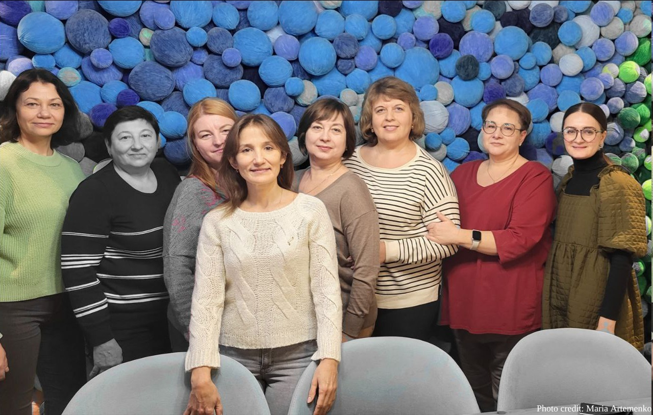 Eight women pose together indoors against a decorative wall featuring blue, purple and green circular patterns, with gray chairs visible in the foreground. Photo credit: Maria Artemenko.
