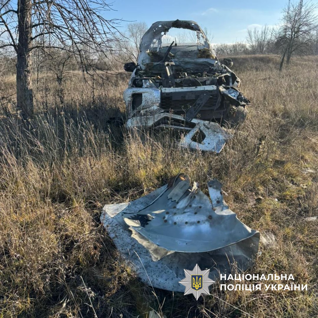 A severely damaged and burned-out vehicle in the Kharkiv Oblast, Ukraine, sits in a bare winter field with dried grass and leafless trees, while a large fragment of charred metal debris lies in the foreground. The wreckage shows extensive fire damage with the vehicle's cabin and front end completely destroyed under a clear blue sky.