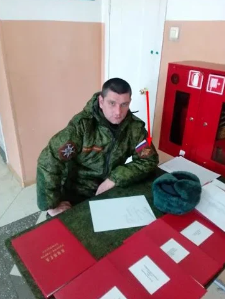 A Russian soldier in camouflage uniform sits at a table with a fur hat placed beside him, reviewing documents and identity cards spread on a red cloth. The setting appears to be an indoor institutional space with beige walls and a red fire safety cabinet visible in the background.