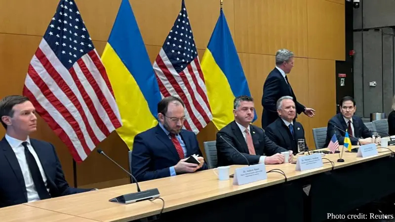 U. S. government officials sit at a conference table during a talks with Ukraine on Russia's war in Geneva, Switzerland, with alternating American and Ukrainian flags displayed behind them against a wood-paneled wall. One person walks behind the seated delegates while others remain seated with documents and microphones on the table, photo credited to Reuters.