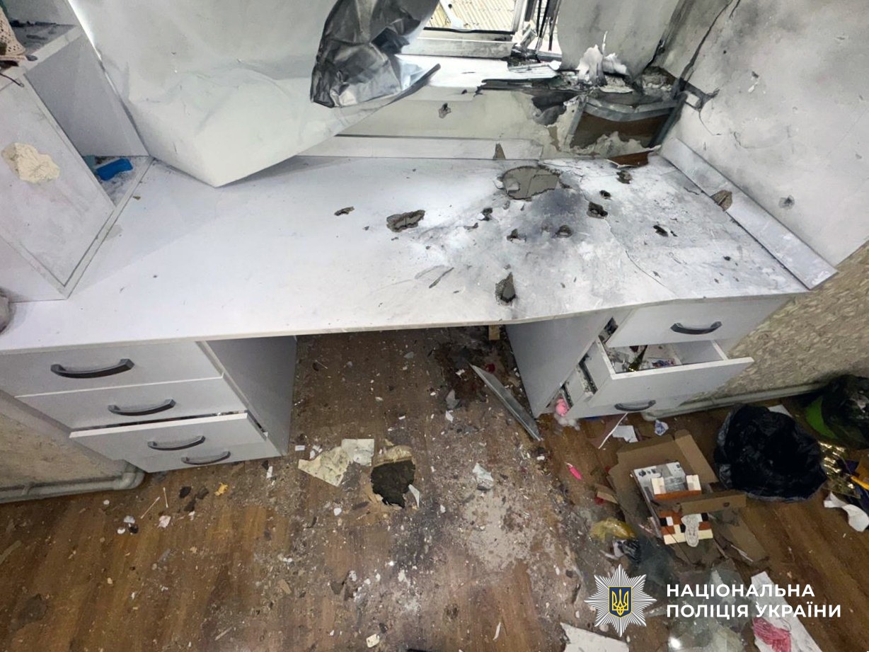 A heavily damaged white desk or workstation shows extensive shrapnel damage across its surface, with drawers pulled open and debris scattered on the floor beneath. The workspace appears to have sustained a blast, with multiple puncture holes visible in the white surface and damaged ceiling elements above.