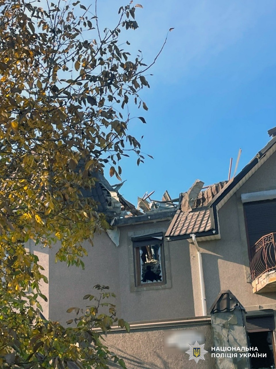 A residential building with severe roof damage showing exposed structural beams and broken tiles against a clear blue sky, with autumn foliage from nearby trees visible in the foreground. The National Police of Ukraine logo appears in the bottom right corner.
