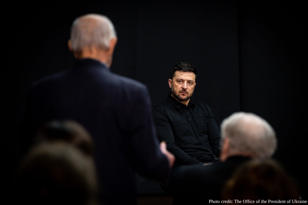 A man in a black shirt sits in focused conversation with others against a dark background, with blurred figures visible in the foreground during what appears to be a formal meeting or interview. Photo credit attributed to The Office of the President of Ukraine.