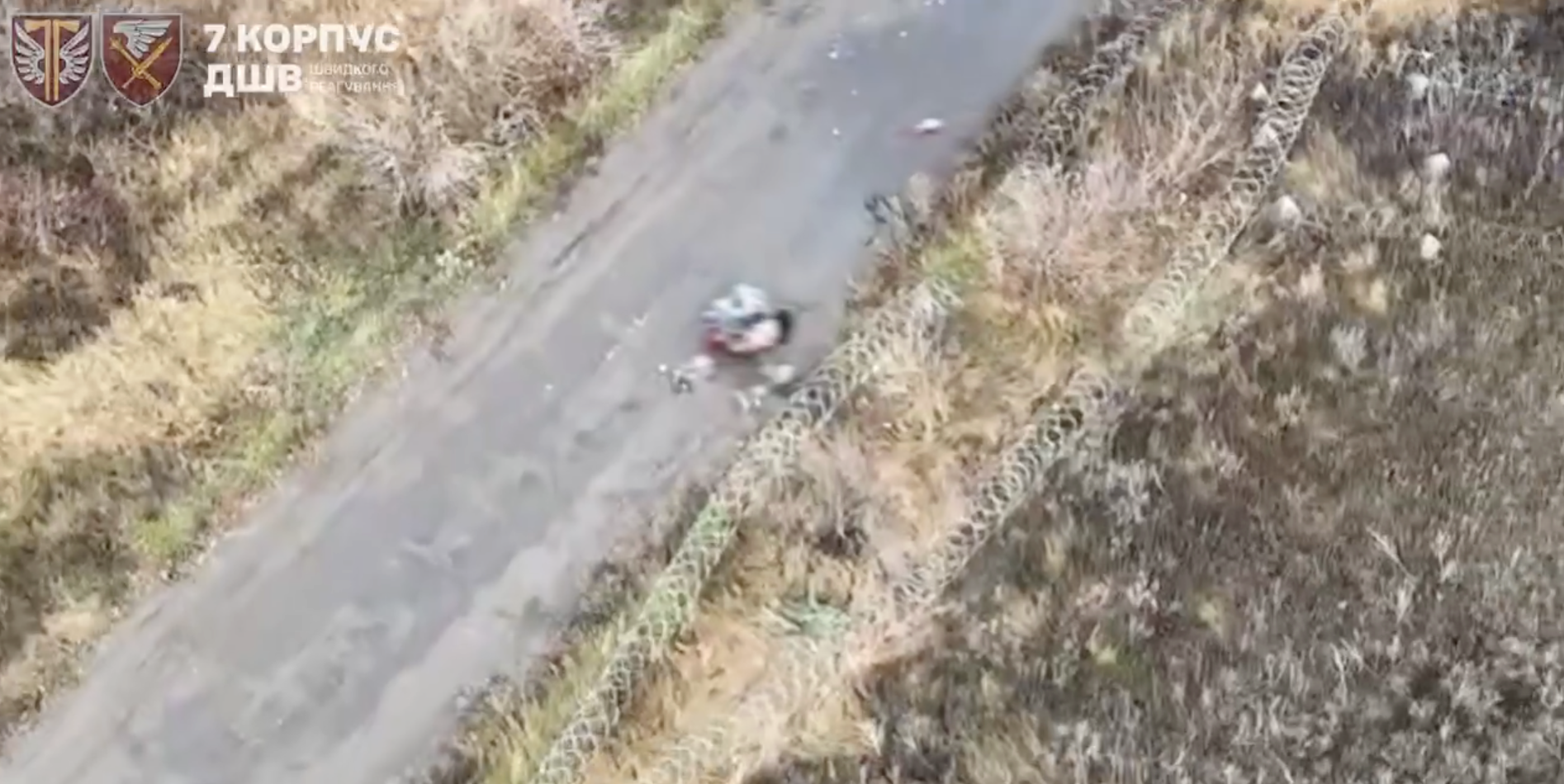 Aerial drone footage shows a damaged or destroyed vehicle on a rural dirt road surrounded by dried vegetation and sparse greenery along the roadside. The grainy overhead view captures the wreckage in the center of the unpaved path with burned or withered fields visible on both sides.