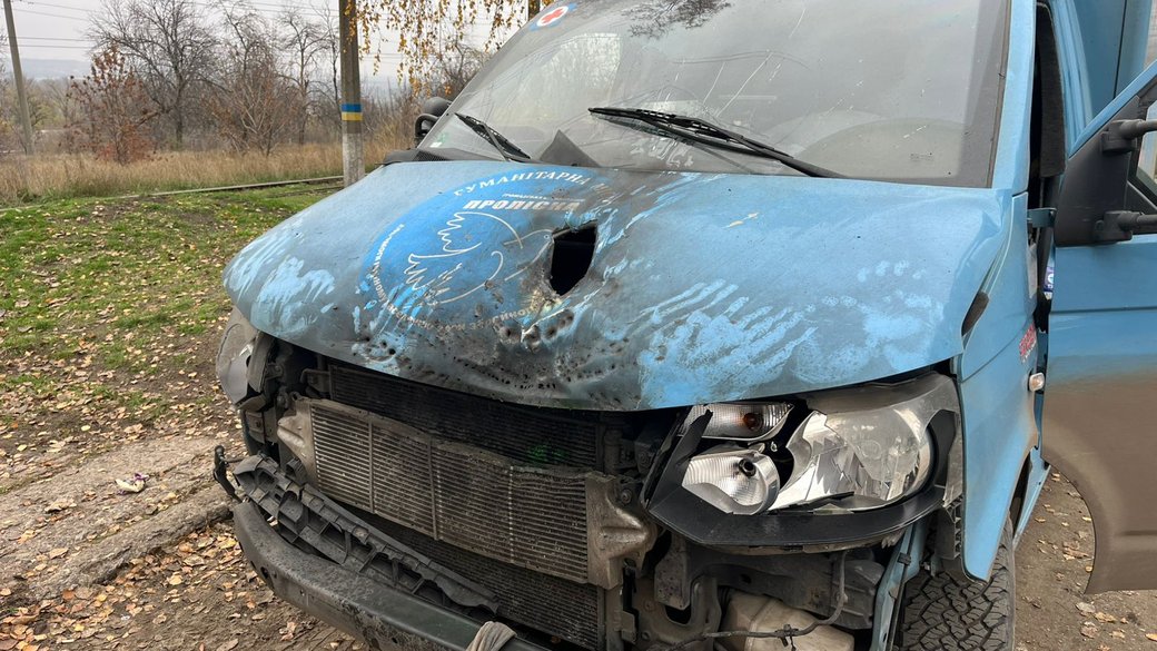 A blue van with significant front-end damage shows a crushed hood with a visible penetration hole, damaged grille, and broken headlight assembly parked on a dirt roadside. The vehicle's weathered and dirty exterior reveals impact damage and paint scrapes, with bare trees and rural landscape visible in the background.