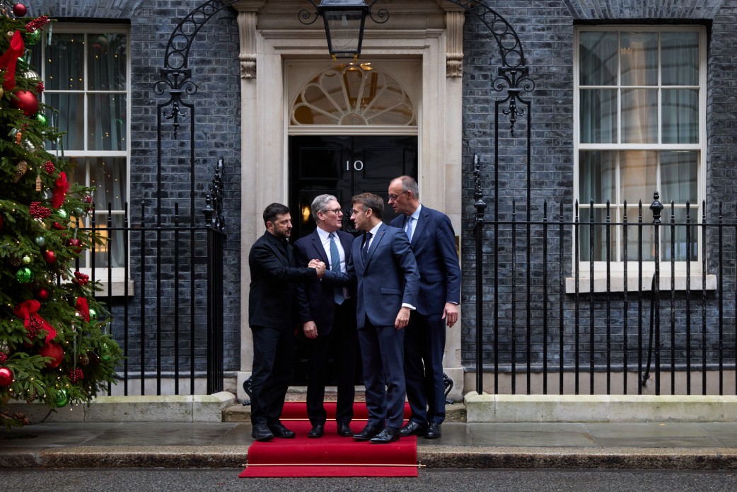 Four men in dark suits shake hands and converse on a red carpet outside the iconic black door of 10 Downing Street, with its distinctive white stone archway and fanlight window. A decorated Christmas tree with red ornaments stands to the left of the Georgian building's entrance.