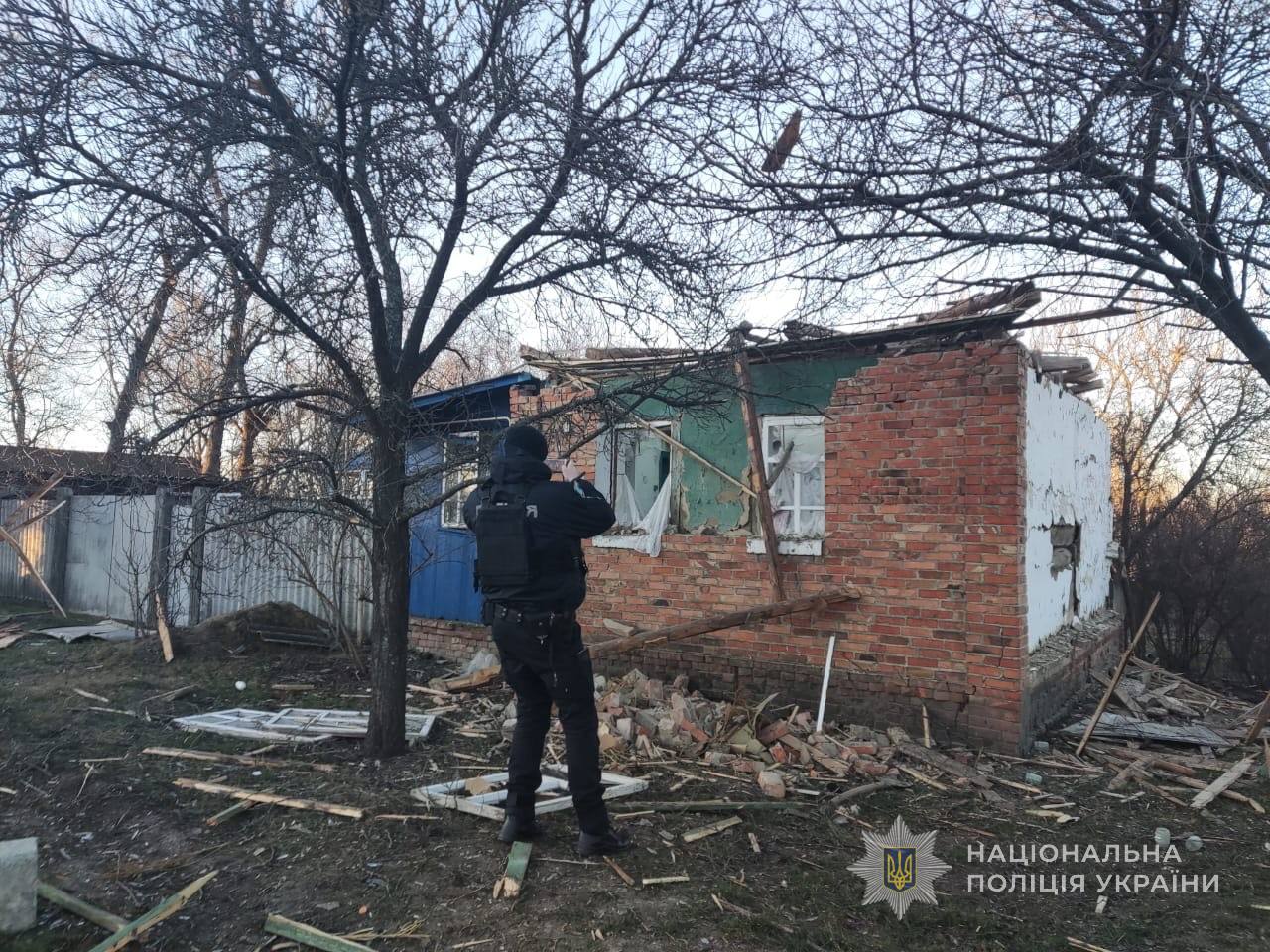 A police officer in dark uniform photographs a severely damaged brick building with its roof destroyed and walls partially collapsed, surrounded by debris and bare trees in what appears to be winter. The National Police of Ukraine logo appears in the bottom right corner of this documentation scene.