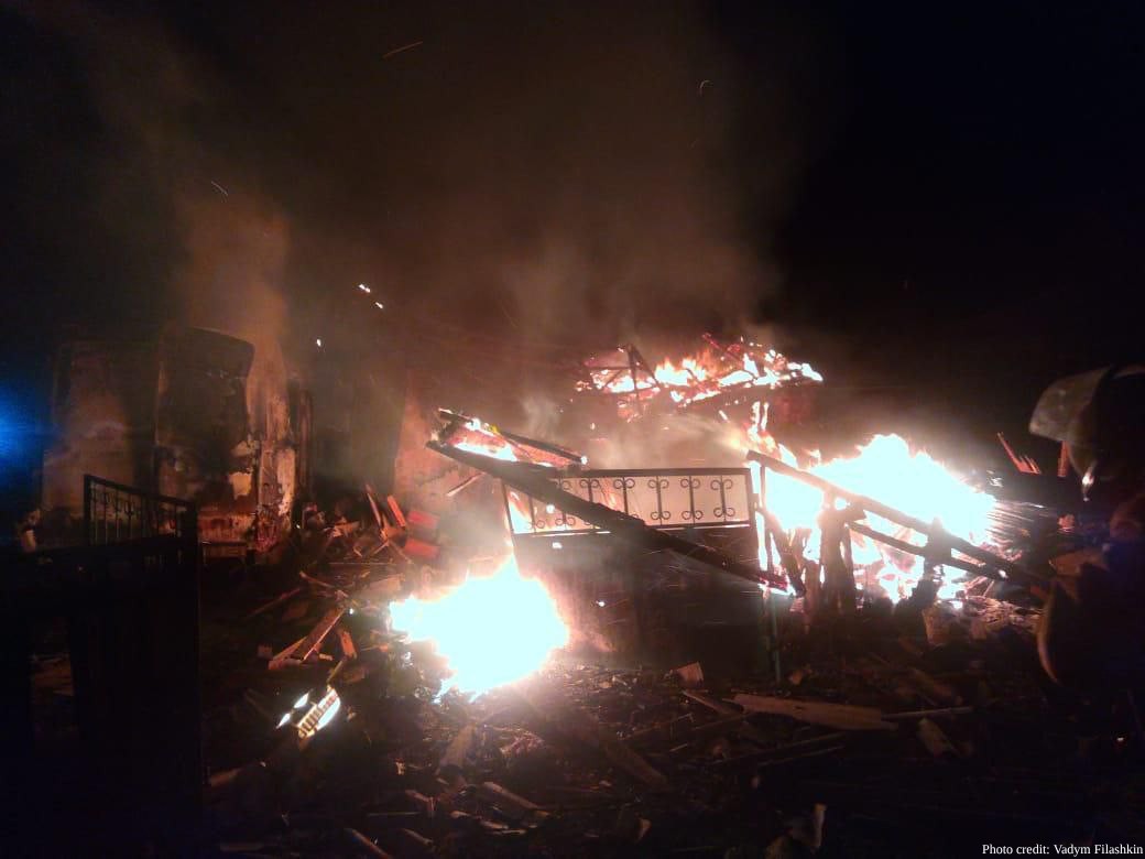 Intense flames engulf the collapsed roof and structural remains of a building at night, with bright orange and blue fire illuminating the debris-scattered ground through thick smoke. A damaged structure with windows is barely visible on the left side of the scene.