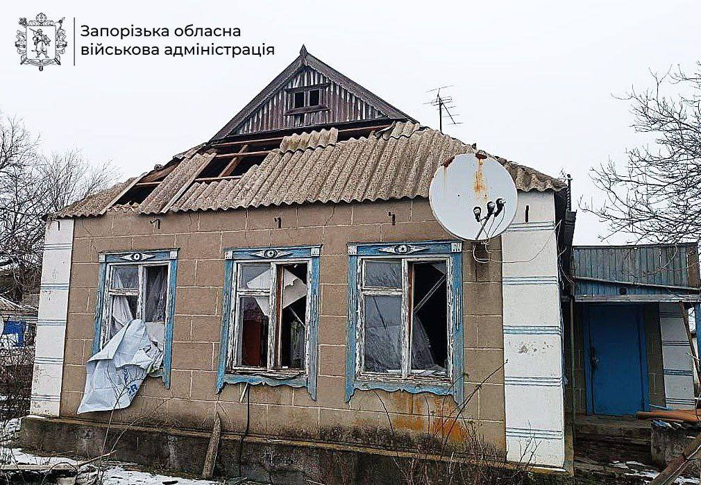 A severely damaged single-story house with a partially collapsed corrugated metal roof, broken blue-trimmed windows with torn curtains, beige block walls, a satellite dish, and a blue door, flanked by bare winter trees under an overcast sky, marked with Zaporizhzhia Regional Military Administration logo.