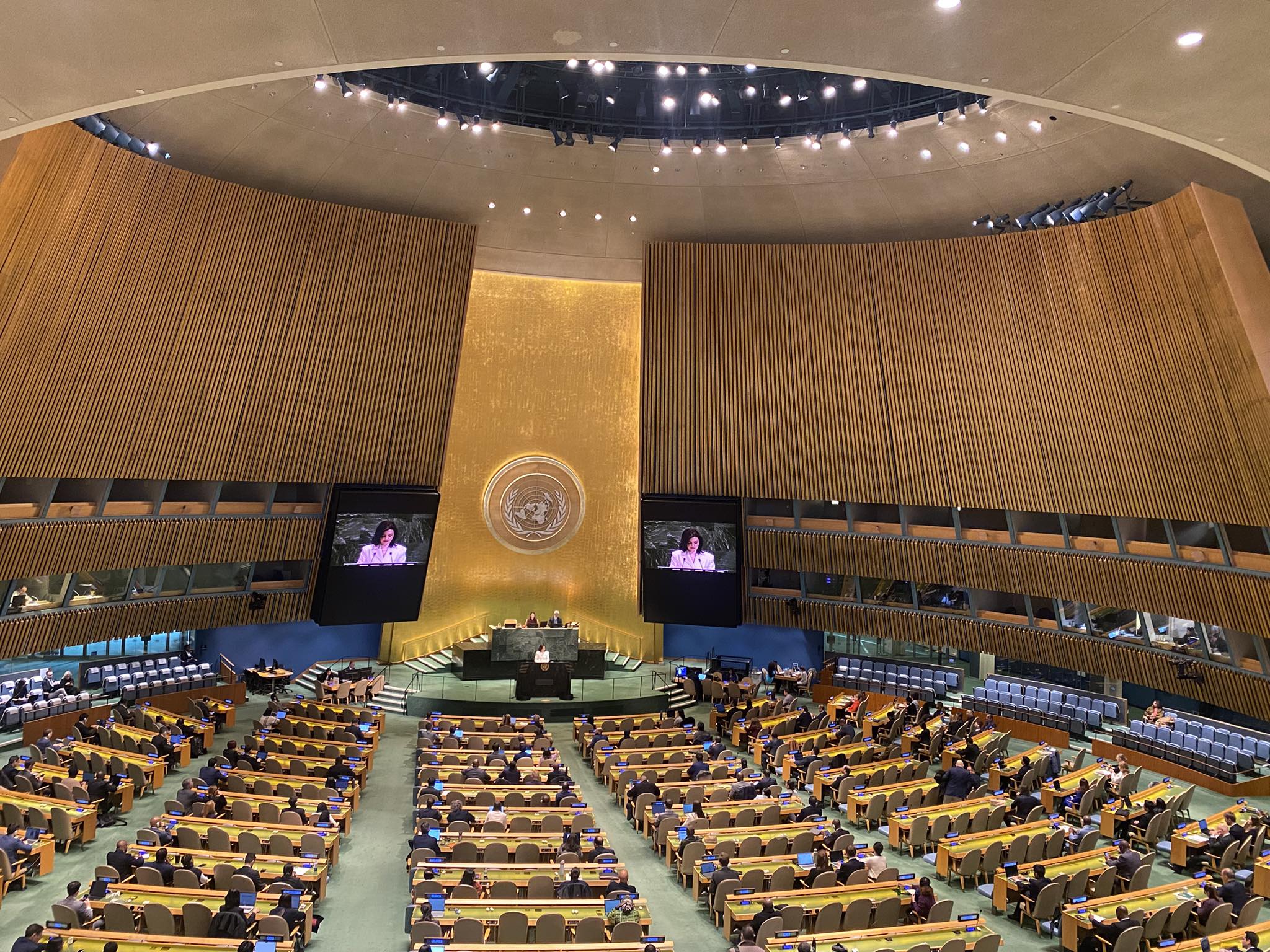 The United Nations General Assembly hall features its distinctive curved wooden wall panels, iconic golden podium with the UN emblem, and tiered seating for delegates arranged in curved rows. The chamber shows an active session in progress with delegates seated at their desks, large video screens displaying a speaker, and the characteristic domed ceiling with its circular opening and recessed lighting.